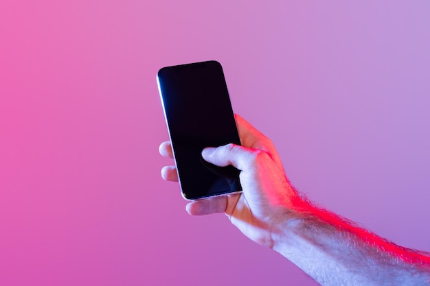 Een persoon houdt een smartphone vast met een donker, niet verlicht scherm tegen een gradiënt paarse achtergrond.