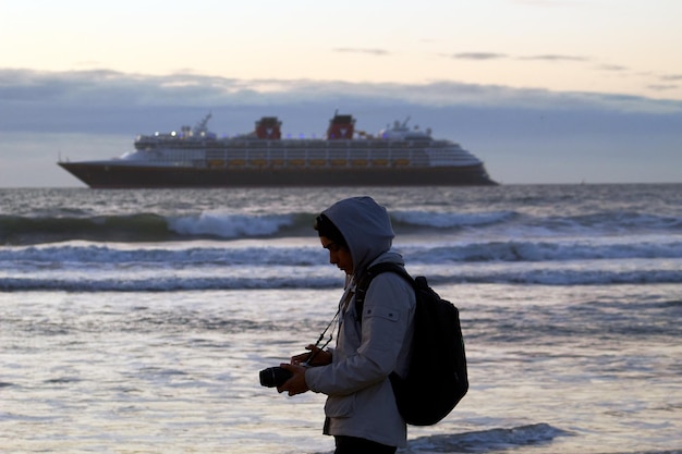 Een persoon met een hoodie staat op het strand voor een cruiseschip.