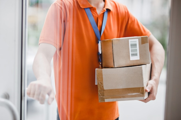 Een persoon met een oranje T-shirt en een naamplaatje staat achter de glazen deur en houdt een deurklink en pakjes voor de klant vast. Vriendelijke werker, bezorgservice van hoge kwaliteit.
