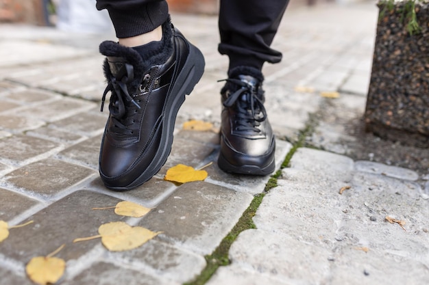 Een persoon met zwarte schoenen en spijkerbroek, lopend op een trottoir, met bladeren op de grond.