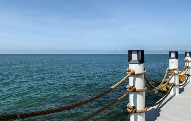 Een pier met een blauwe oceaan en een witte paal met een touw waarop staat 'de zee'