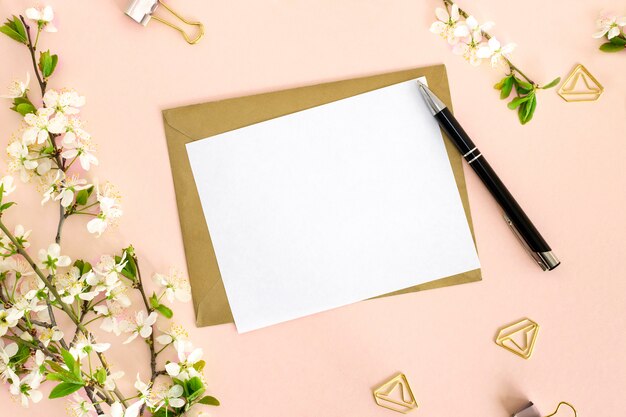 Een platliggende compositie met een mock-up van een witte blanco kaart voor tekst, een envelop van kraftpapier, een pen, kantoorclips en een tak van kers met bloemen op een roze achtergrond. bovenaanzicht.