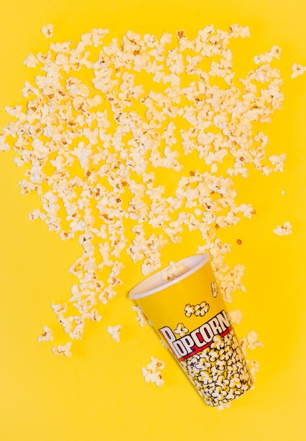 een popcornemmer omgevallen omringd door veel popcorn met een gele achtergrond