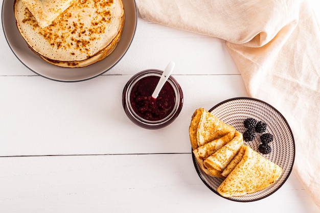 Een portie vers gebakken pannenkoeken op een bord met bessen en een potje raspberry jam op een witte houten tafel Het concept van ontbijt op Maslenitsa