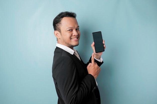 Een portret van een gelukkige Aziatische zakenman lacht en houdt zijn smartphone vast met kopieerruimte op het it39s-scherm in een zwart pak geïsoleerd door een blauwe achtergrond
