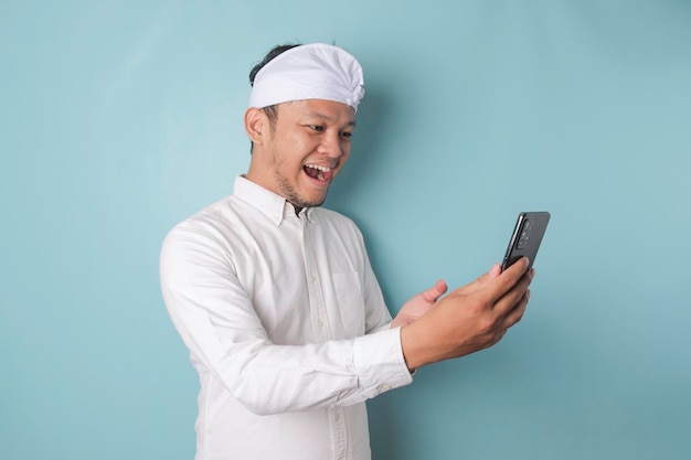 Een portret van een gelukkige Balinese man lacht en houdt zijn smartphone vast met udeng of traditionele hoofdband en wit overhemd geïsoleerd door een blauwe achtergrond