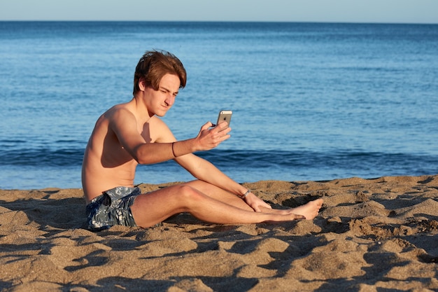 Een portret van een jonge, geschikte blanke man die zit en een selfie maakt op het strand