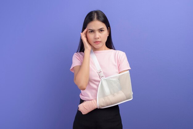 Een portret van een jonge vrouw gestrest en gefrustreerd met een gewonde arm in een mitella over een blauwe achtergrond in studio, verzekering en gezondheidszorgconcept