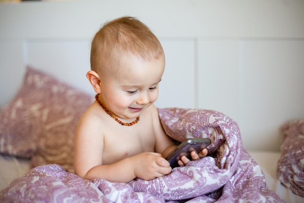 Een portret van een schattige baby die smartphone speelt, liggend in een bed onder een dekbed Peuterjongen met een amberkleurige ketting met mobiele telefoon