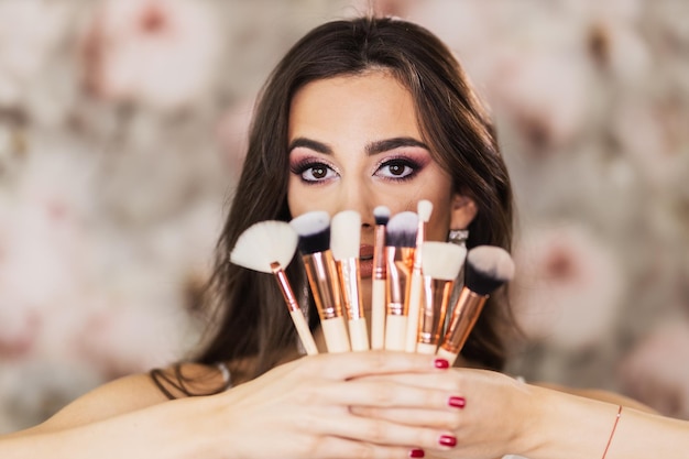 Een portret van een schattige jonge vrouw met mooie ogen, die make-upborstels vasthoudt en een mooie make-up op haar gezicht vertegenwoordigt.