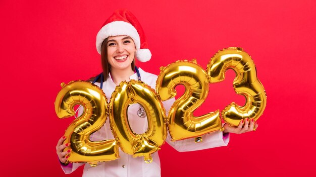 Een portret van een vrolijke verpleegster in een witte jas en een kerstmanhoed houdt de figuren van het nieuwe jaar 2023 vast van ballonnen op een rode achtergrond