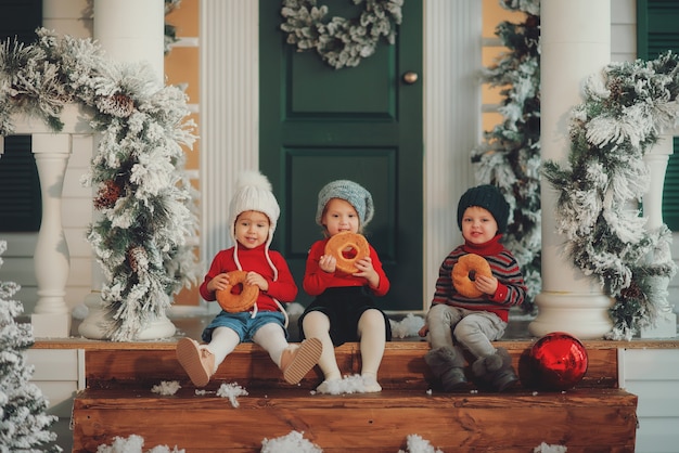 Een portret van kinderen die samen op de veranda van hun huis zitten, Eat bagels. Vrolijk kerstfeest gelukkig nieuwjaar. Werf met een kerstboom, lichtjes en versieringen. Wondertijd.