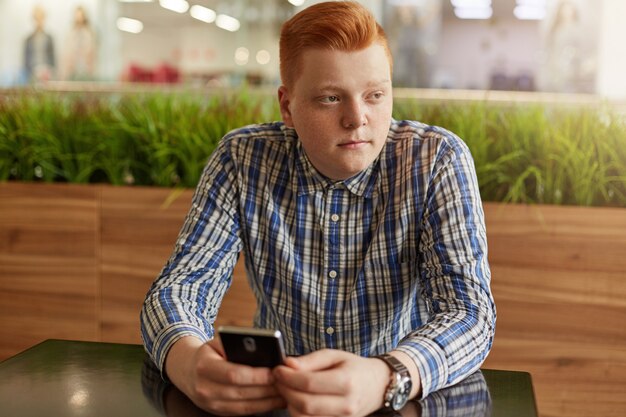 Een portret van nadenkende tiener met rood haar en sproeten die gecontroleerd overhemd dragen die mobiele telefoonzitting houden bij houten lijst