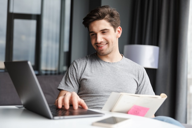 een positieve optimistische jonge man binnenshuis met behulp van laptopcomputer met documenten.