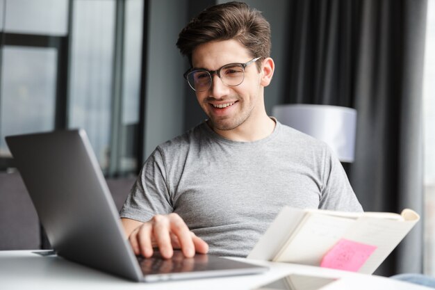 een positieve optimistische jonge man binnenshuis met behulp van laptopcomputer met documenten.