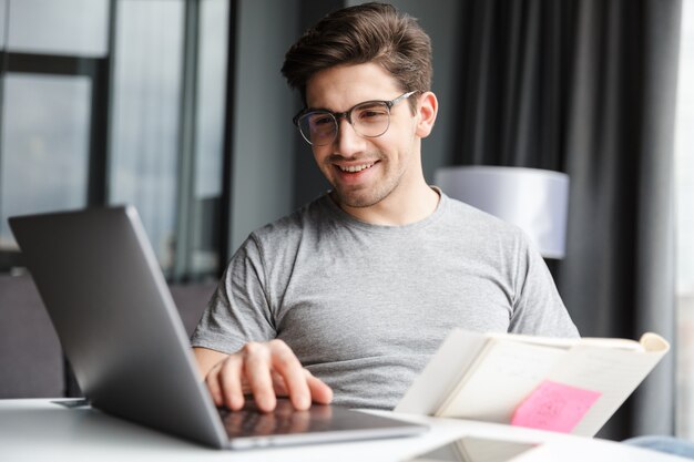 Een positieve optimistische jonge man binnenshuis met behulp van laptopcomputer met documenten.