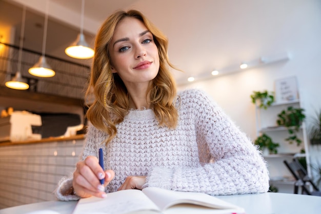 een positieve optimistische mooie jonge blonde vrouw zit in café binnenshuis en bestudeert het schrijven van notities.