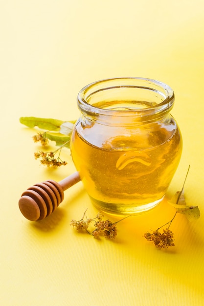 Een pot vloeibare honing van lindebloemen en een stok met honing op een geel oppervlak.
