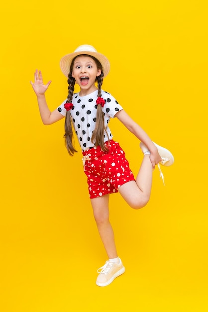 Een prachtig klein meisje speelt in een strooien hoed en korte broek Een mooi kind geniet van de zomer en glimlacht breed en speelt wat rond op een gele geïsoleerde achtergrond