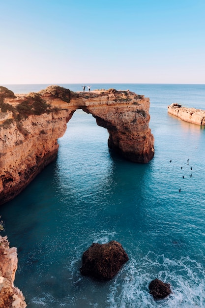 Een prachtige formatieboog van rotsen met een paar erop, enkele vliegende vogels en een golf die bij de zonsopgang tegen een rots in de algarve, portugal beukt. kopieer ruimte.