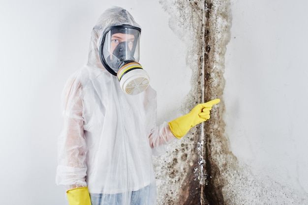 Een professionele desinfector in overall en een masker wijst met de vinger naar de zwarte schimmel aan de muur. verwijdering van de schimmel in het appartement en huis. aspergillus.