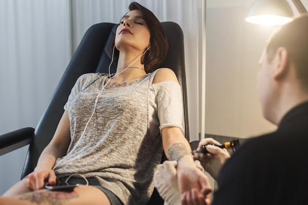 Foto een professionele tatoeëerder die een foto van de vrouw maakt met de machine terwijl het meisje naar muziek luistert. tattoo kunst op het lichaam. apparatuur voor het maken van zwarte inkt van een jartattoo-kunst.