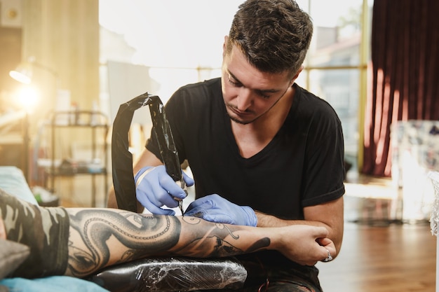 Een professionele tatoeëerder die tatoeage op de arm van een jonge man doet door een machine met zwarte inkt