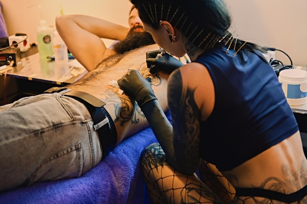 Een professionele vrouwelijke tatoeëerder maakt zwart-witte tatoeage op bebaarde mannelijke torso in een salon.