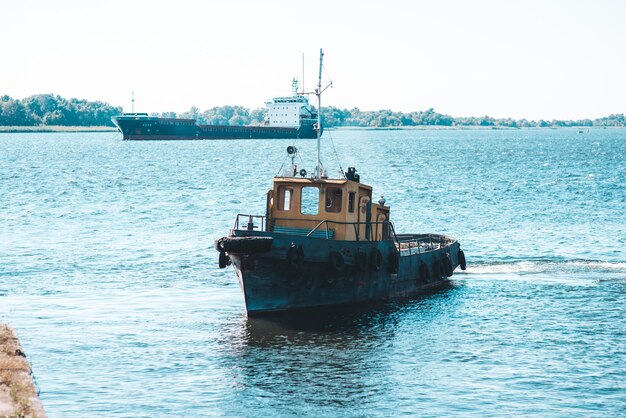 Een riviersleepboot komt de haven binnen tegen de achtergrond van een bulkcarrier in de rede