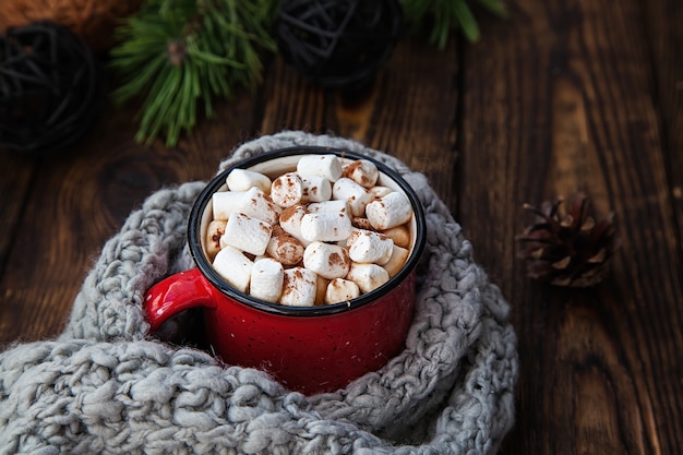 Een rode beker met warme drank met marshmallow en gebreide sjaal op houten kerst