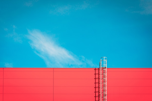 Een rode moderne gevel van een industrieel gebouw, pakhuis of winkelcentrum met een lange metalen trap tegen de blauwe lucht