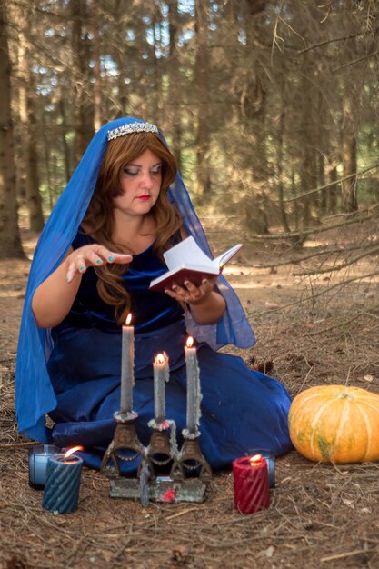 Een roodharige heks in een blauwe jurk en een sluier op haar hoofd in het bos leest spreuken uit een boek over kaarsen