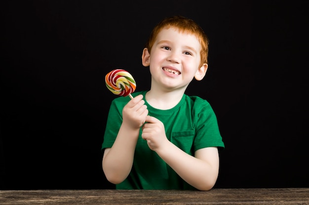 Een roodharige jongen met een mooi gezicht eet een zoet veelkleurig snoepje op een stokje, een jongen met een lolly gemaakt van suiker