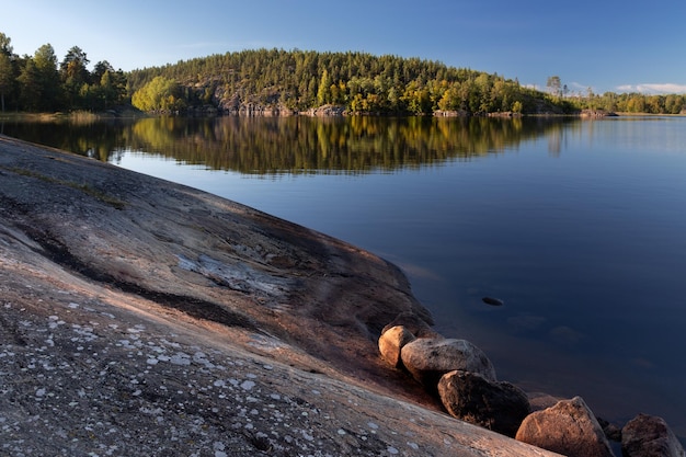 Een rotsachtige helling aan de kust van het meer en eiland met dichte bomen weerspiegeld in meerwater Karelië Rusland