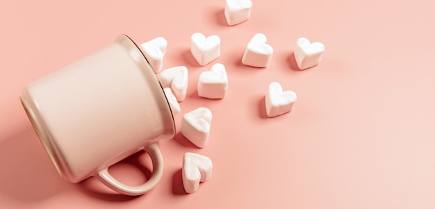 Een roze mok ligt op zijn kant tegen een roze oppervlak, lichtroze marshmallows worden er in de vorm van harten vanaf gestrooid
