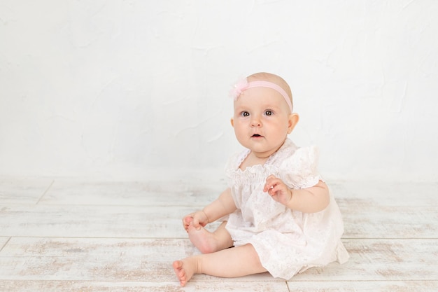 Een schattig babymeisje in een witte jurk zit zes maanden op de grond in een lichte kamer, een plek en ruimte voor tekst