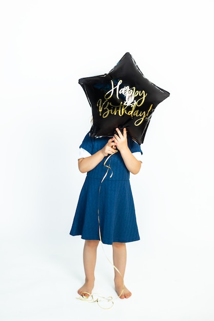 Een schattig blank babymeisje bedekt haar gezicht met een zwarte gelballon met de tekst Happy Birthday. Foto in studio tegen een witte achtergrond