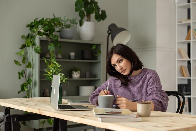 een schattig brunette meisje in een lila trui zit aan een tafel met een laptop die aantekeningen maakt