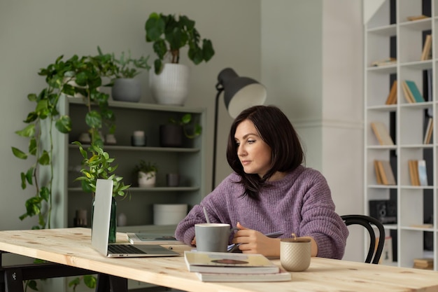 een schattig brunette meisje in een lila trui zit aan een tafel met een laptop