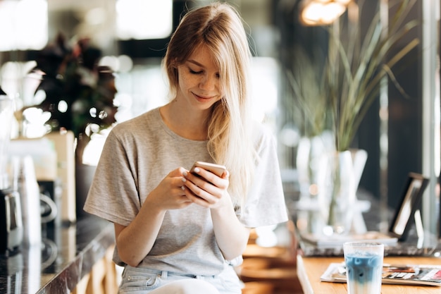 Een schattig dun blond meisje, gekleed in casual stijl, drinkt koffie en kijkt naar haar telefoon in een coffeeshop. .