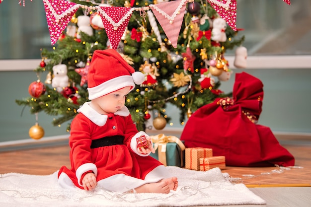 Een schattig kind verkleed als kerstman speelt in de buurt van een versierde kerstboom in de woonkamer.