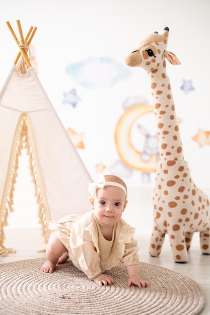 Een schattig klein Europees meisje met bruine ogen in een beige bodysuit gemaakt van natuurlijke stof speelt thuis in de kinderkamer tegen de achtergrond van een wigwam en knuffels