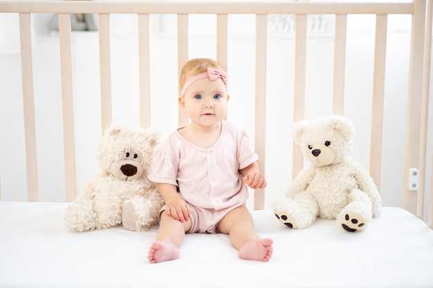 Een schattig klein gezond meisje tot een jaar oud in een roze bodysuit gemaakt van natuurlijke stof zit in een wieg met teddyberen op wit beddengoed in de slaapkamer kijkend naar de camera de baby is thuis