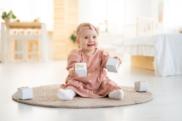 Een schattig klein gezond meisje tot een jaar oud in een roze jurk gemaakt van natuurlijke stof zit op een kleed in de woonkamer van het huis te spelen met houten blokjes de ontwikkeling van het kind thuis