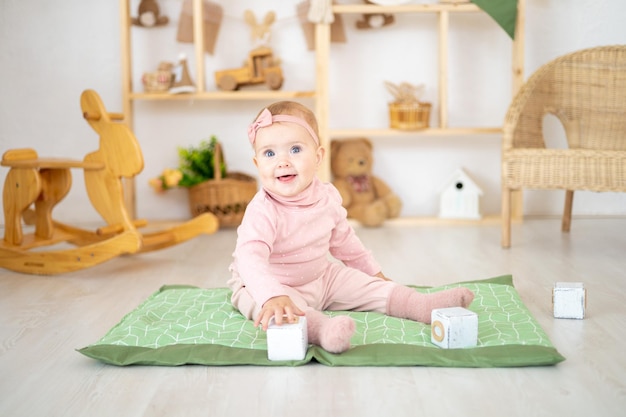 Een schattig klein gezond meisje tot een jaar oud in een roze pak gemaakt van natuurlijke stof zit op een tapijt in een kinderkamer met houten educatief speelgoed glimlachend naar de camera kijkend