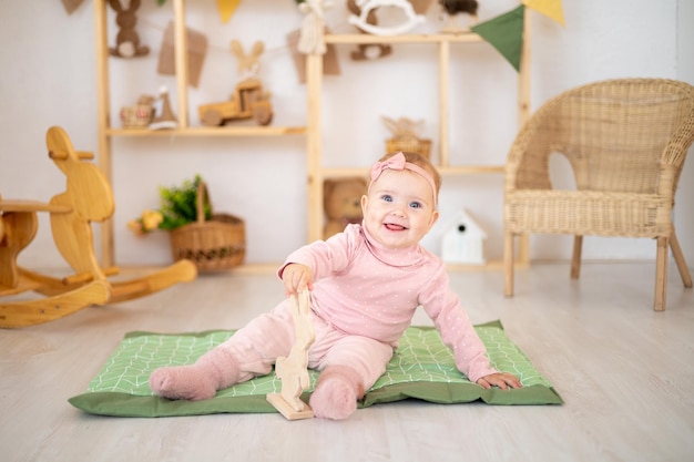 Een schattig klein gezond meisje tot een jaar oud in een roze pak gemaakt van natuurlijke stof zit op een tapijt in een kinderkamer met houten educatief speelgoed glimlachend naar de camera kijkend