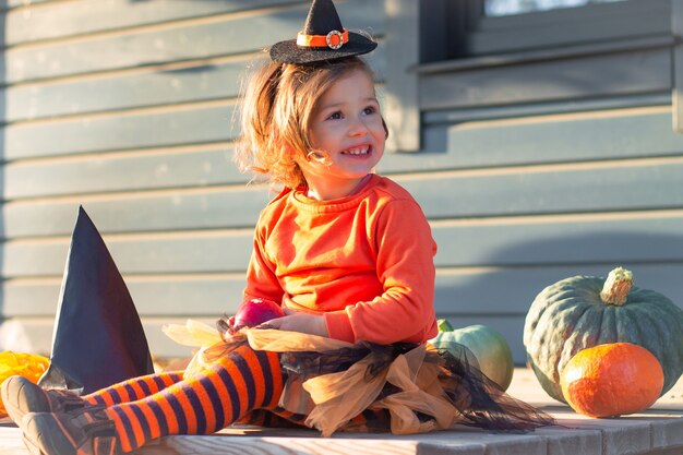 Een schattig klein meisje 2-3 in een oranje en zwart heksenkostuum zit naast pompoenen op het terras van een houten grijs huis