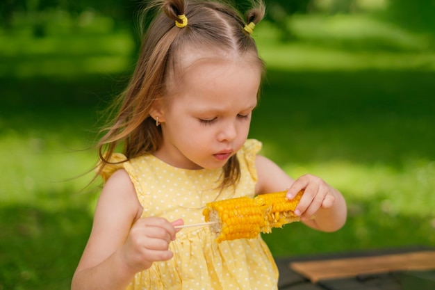 Een schattig klein meisje eet gekookte maïs zittend op een bankje in het park