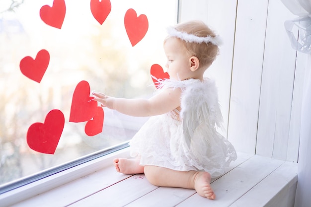 Een schattig klein meisje in een engelenkostuum met vleugels zit op de vensterbank bij het raam tegen een achtergrond van rode harten Valentijnsdag concept Valentijnsdag