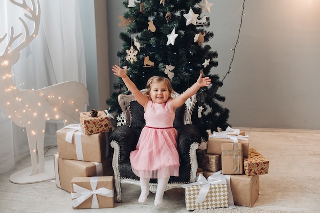 Een schattig klein meisje in een jurk zit omringd door vele dozen met kerstcadeaus en wil ze openen